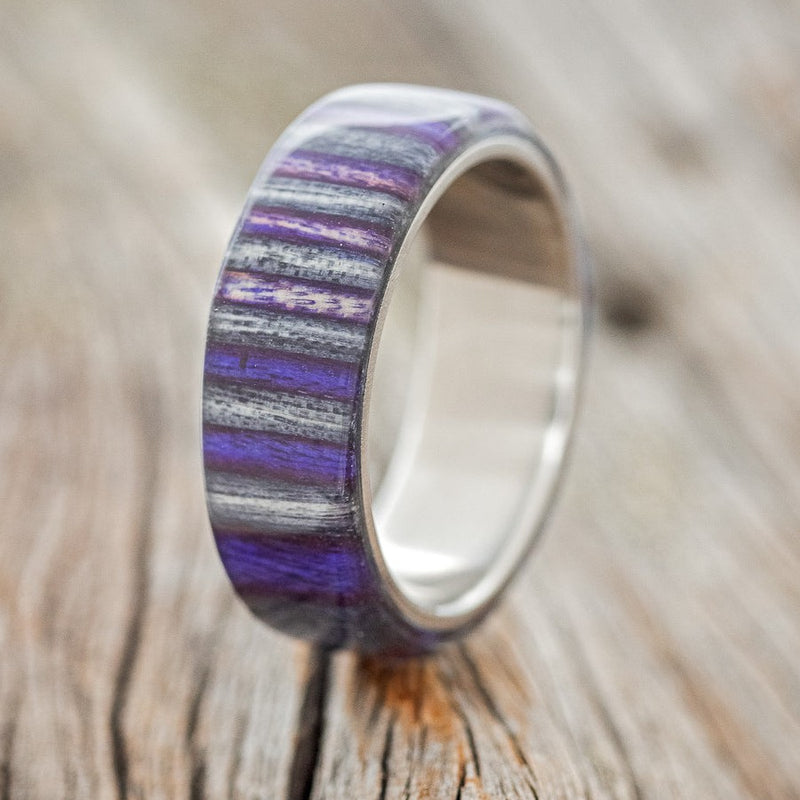 Shown here is "Haven", a custom, handcrafted men's wedding ring featuring birch wood overlay that has been dyed purple and grey, upright facing left. Additional overlay options are available upon request.