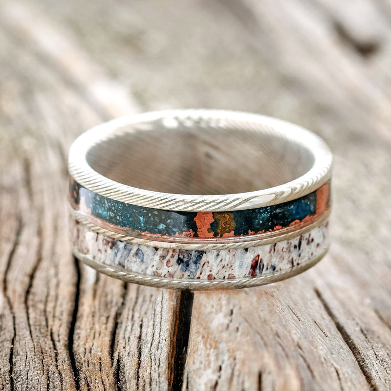 Shown here is "Dyad" is a custom, handcrafted men's wedding ring featuring 2 channels with patina copper and antler inlays, laying flat.