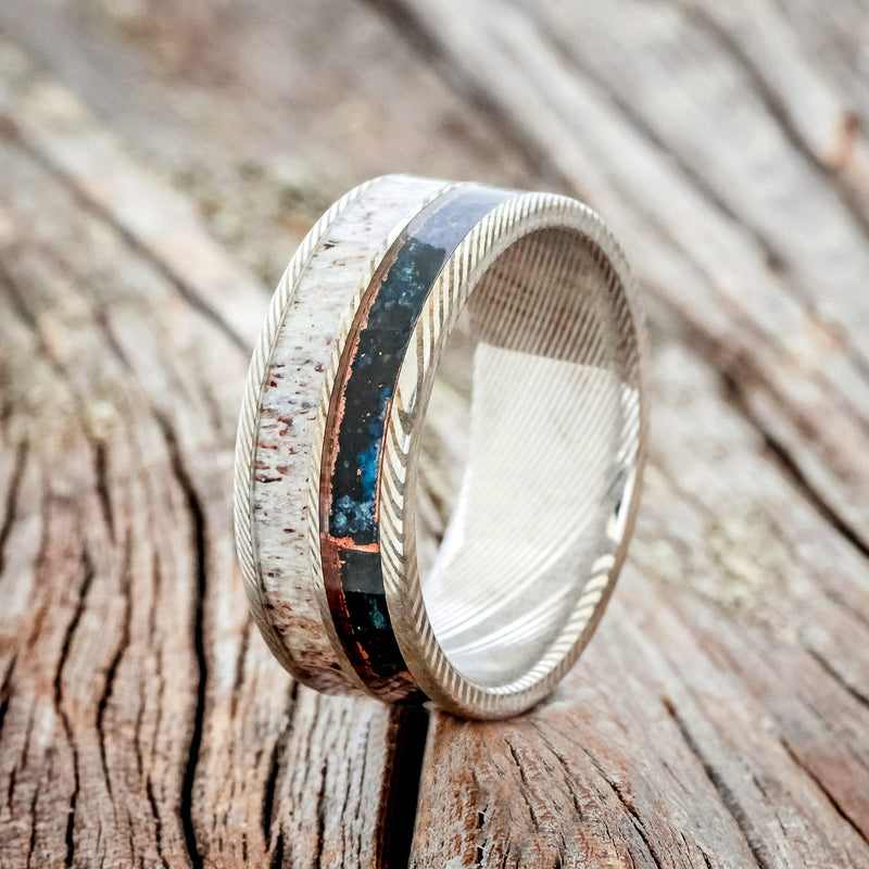 Shown here is "Dyad" is a custom, handcrafted men's wedding ring featuring 2 channels with patina copper and antler inlays, upright facing left.