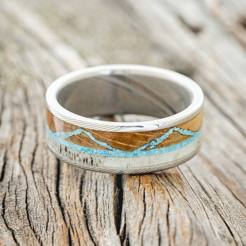Shown here is "The Expedition", a custom, handcrafted men's wedding ring featuring a mountain engraving with whiskey barrel oak, antler and turquoise inlays, laying flat. Additional inlay options are available upon request.