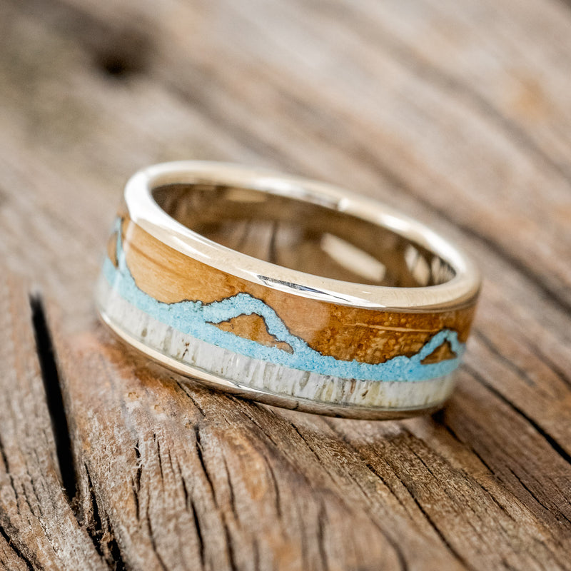 Shown here is "The Expedition", a custom, handcrafted men's wedding ring featuring a mountain engraving with whiskey barrel oak, antler and turquoise inlays, tilted left. Additional inlay options are available upon request.