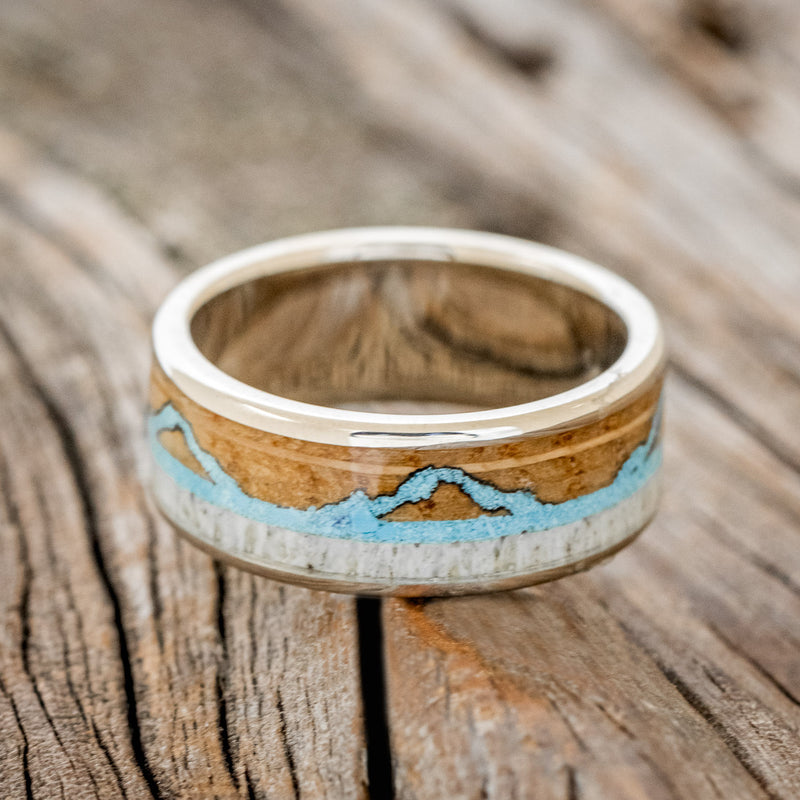 Shown here is "The Expedition", a custom, handcrafted men's wedding ring featuring a mountain engraving with whiskey barrel oak, antler and turquoise inlays, laying flat. Additional inlay options are available upon request.