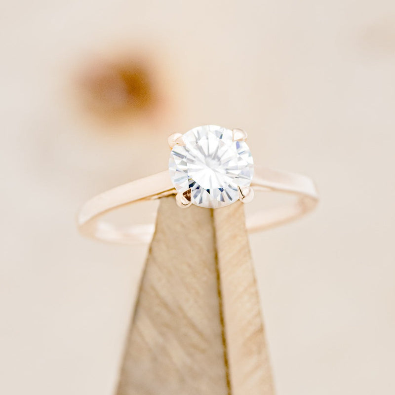 Shown here is a solitaire-style moissanite women's engagement ring with 4 claw prongs, on stand front facing. Many other center stone options are available upon request. 
