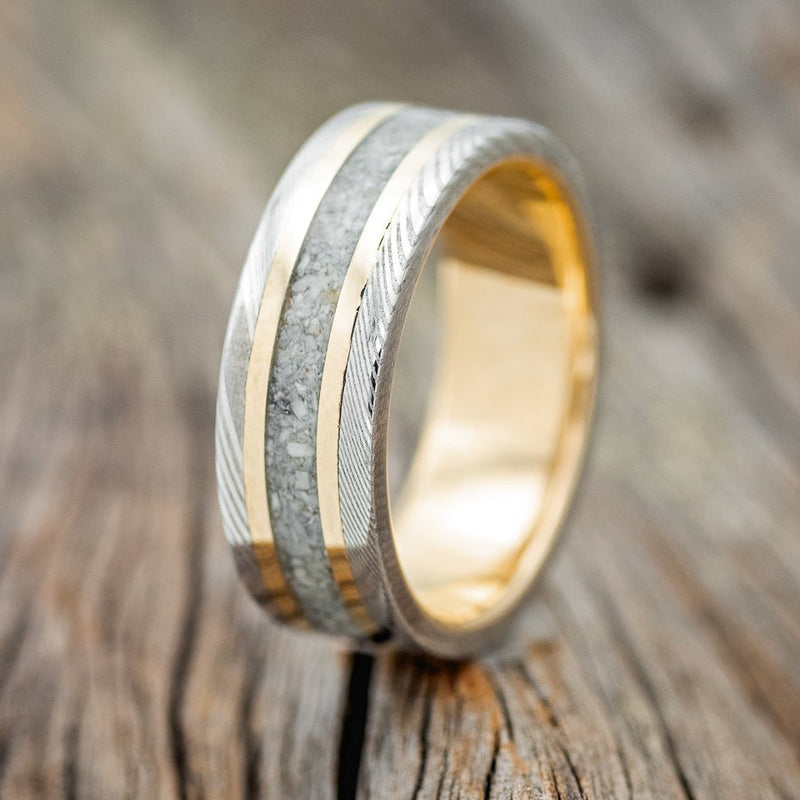 Shown here is "Kalder", a custom, handcrafted men's wedding ring featuring a 14K yellow gold band with 2 14K yellow gold inlays, a centered elk tooth inlay, and Damascus steel overlay, upright facing left. Additional inlay options are available upon request.