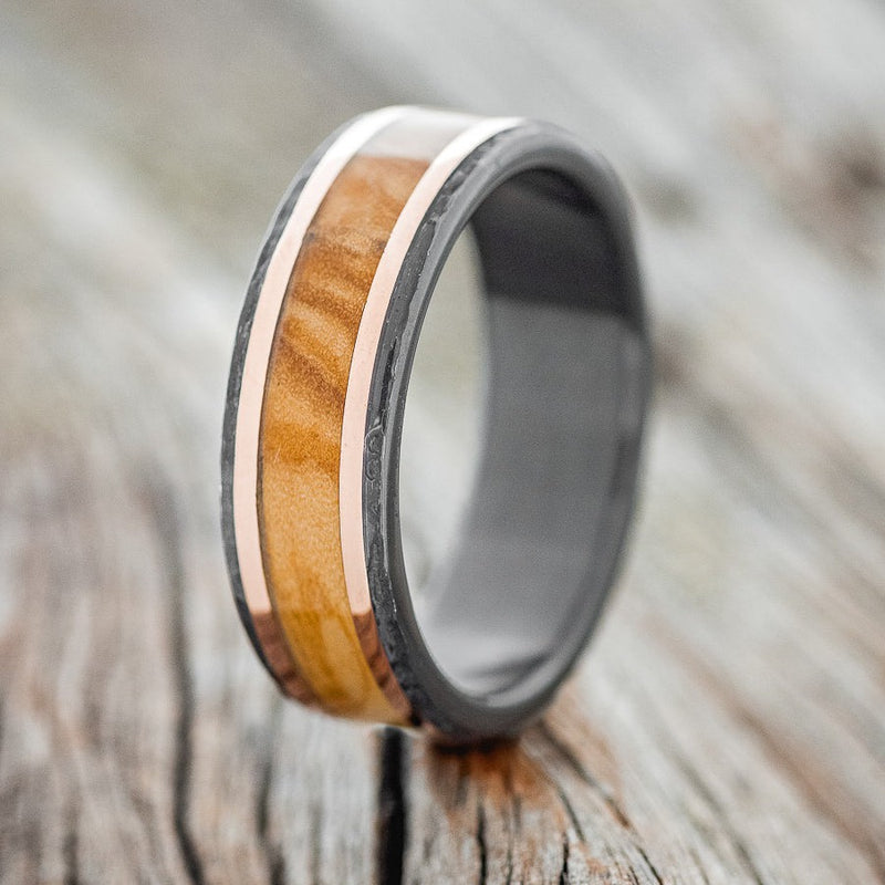 Shown here is "Hollis", a custom, handcrafted men's wedding ring featuring Bethlehem olive wood & 14K rose gold inlays on a hammered, fire-treated black zirconium band, upright facing left. Additional inlay options are available upon request.