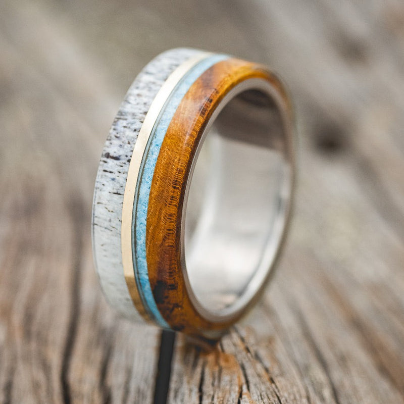 Shown here is "Banner", a custom, handcrafted men's wedding ring featuring turquoise,  ironwood, naturally shed antler inlays, and a 14K yellow gold inlay, upright facing left. Additional inlay options are available upon request.