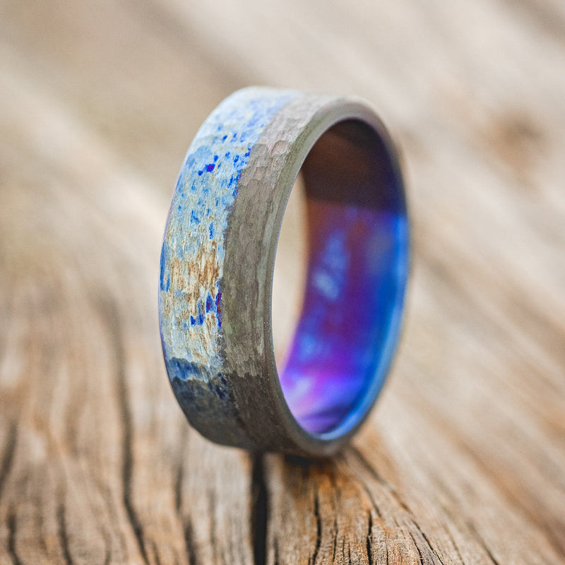 Shown here is a custom, handcrafted wedding band featuring a fire-treated titanium ring with a black zirconium overlay and a hammered finish, upright facing left. Additional inlay options are available upon request.