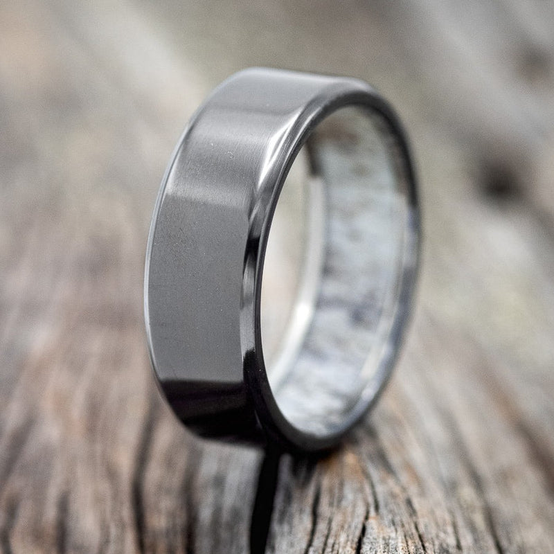 Shown here is a custom, handcrafted men's wedding ring featuring a unique antler lining shown here on a fire-treated black zirconium band, upright facing left. Additional inlay options are available upon request.