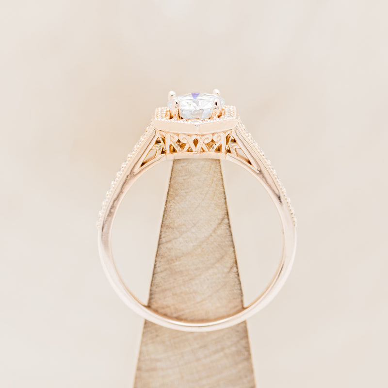 Shown here is "Odessa", a geometric-style moissanite women's engagement ring with a diamond halo and diamond accents, side view on stand. Many other center stone options are available upon request.