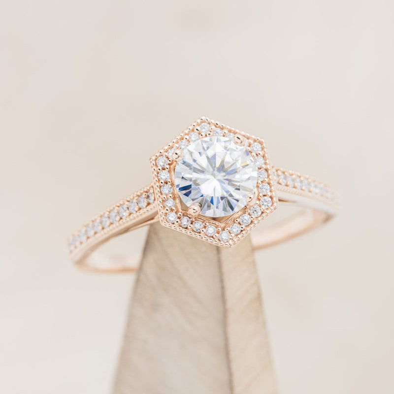 Shown here is "Odessa", a geometric-style moissanite women's engagement ring with a diamond halo and diamond accents, on stand front facing. Many other center stone options are available upon request.