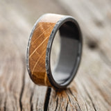 Shown here is "Rainier", a custom, handcrafted men's wedding ring featuring whiskey barrel wood inlay, shown here on a fire-treated black zirconium band with a hammered finish, upright facing left. Additional inlay options are available upon request.