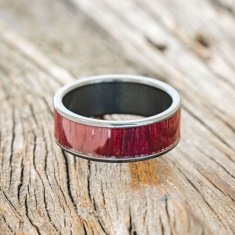 Shown here is "Rainier", a custom, handcrafted men's wedding ring featuring purpleheart wood inlay on a fire-treated black zirconium band, laying flat. Additional inlay options are available upon request.