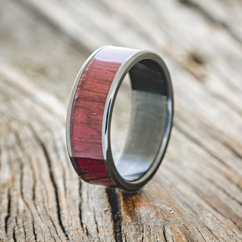 Shown here is "Rainier", a custom, handcrafted men's wedding ring featuring purpleheart wood inlay on a fire-treated black zirconium band, upright facing left. Additional inlay options are available upon request.