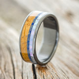 Shown here is "Rainier", a custom, handcrafted men's wedding ring featuring a mother of pearl, lapis lazuli, and whiskey barrel oak inlays on a fire-treated black zirconium band, upright facing left. Additional inlay options are available upon request.