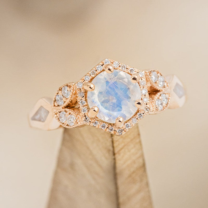 The "Lucy in the Sky" petite is a halo-style, faceted moonstone women's engagement ring with a diamond accents and diamond dust inlays, on stand facing slightly right. Many other center stone options are available upon request.