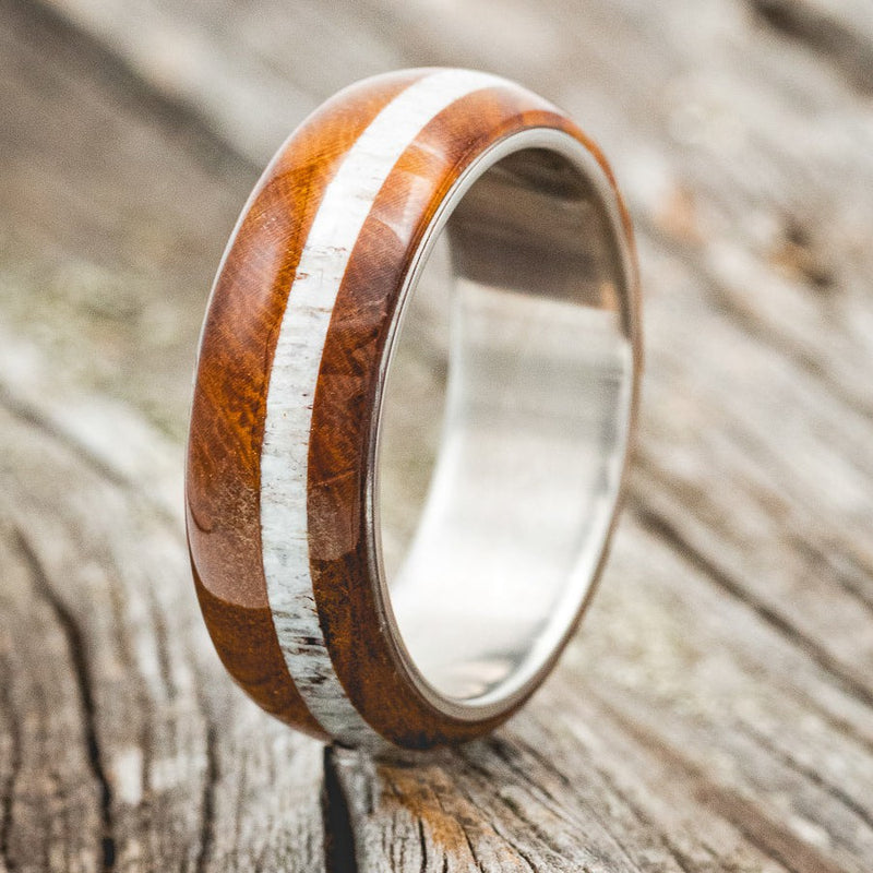 Shown here is "Remmy", a custom, handcrafted men's wedding ring featuring an ironwood overlay and an antler inlay, upright facing left. Additional inlay options are available upon request.
