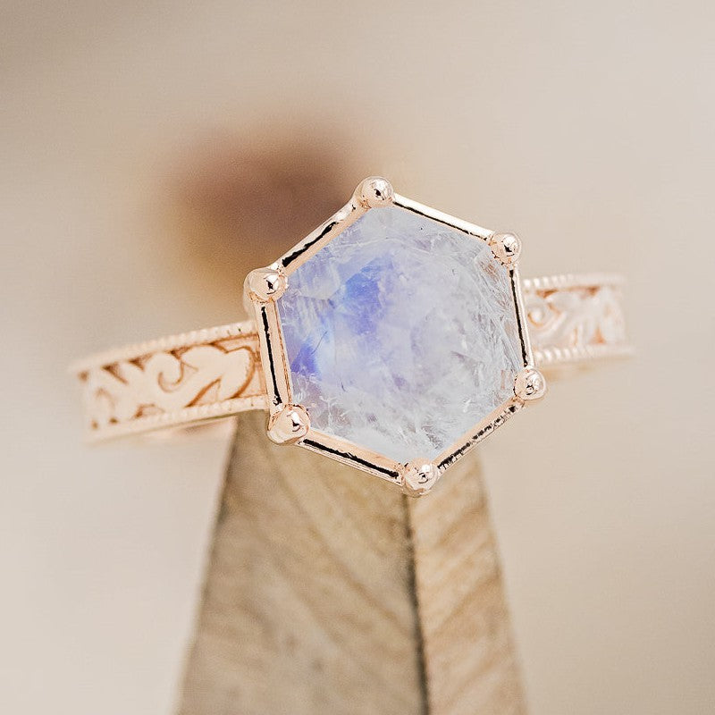 Shown here is "Hey There Delilah", a floral-style hexagon moonstone women's engagement ring with a floral engraved band, on stand facing slightly right. Many other center stone options are available upon request.