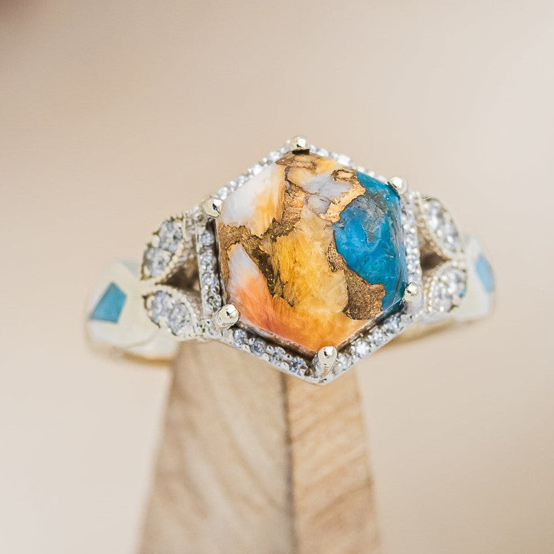 Shown here is "Lucy in the Sky", a halo-style hexagon spiny oyster turquoise women's engagement ring with diamond accents and turquoise inlays, on stand front facing. Many other center stone options are available upon request.