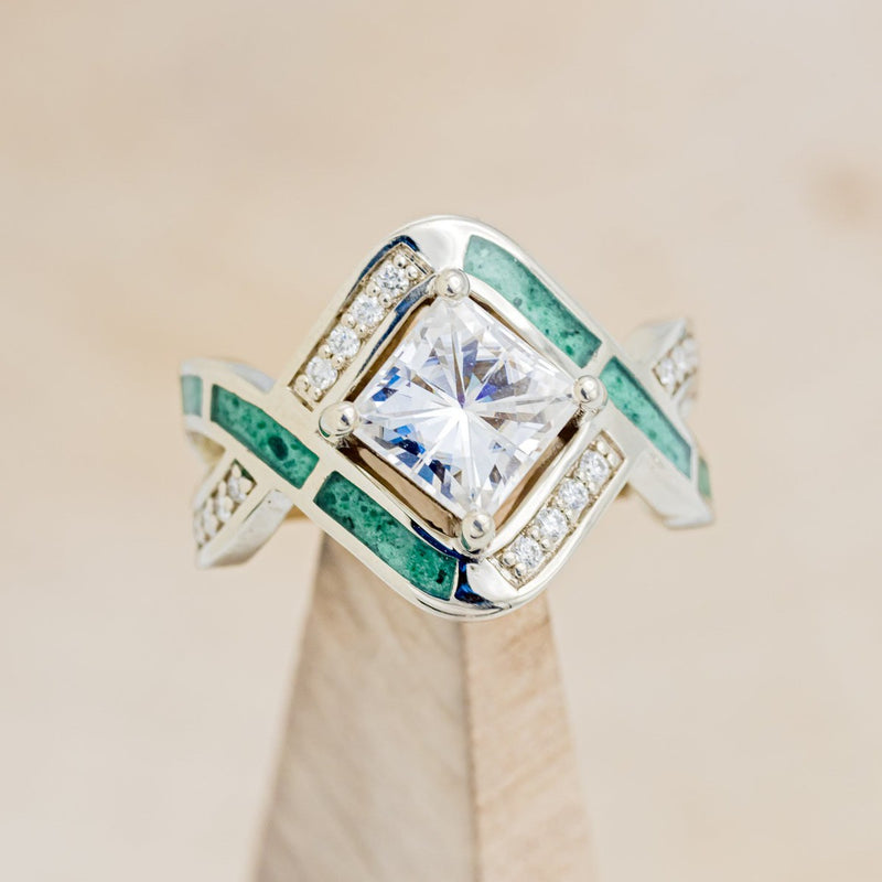 Shown here is "Helix", a geometric-style princess cut moissanite women's engagement ring with diamond accents and malachite inlays, on stand front facing. Many other center stone options are available upon request. 
