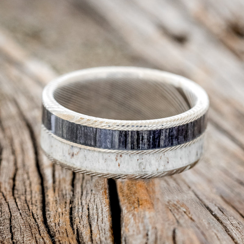 Shown here is "Dyad", a custom, handcrafted men's wedding ring featuring 2 channels with grey birch wood and antler inlays, shown here on an etched Damascus Steel band, laying flat. Additional inlay options are available upon request.