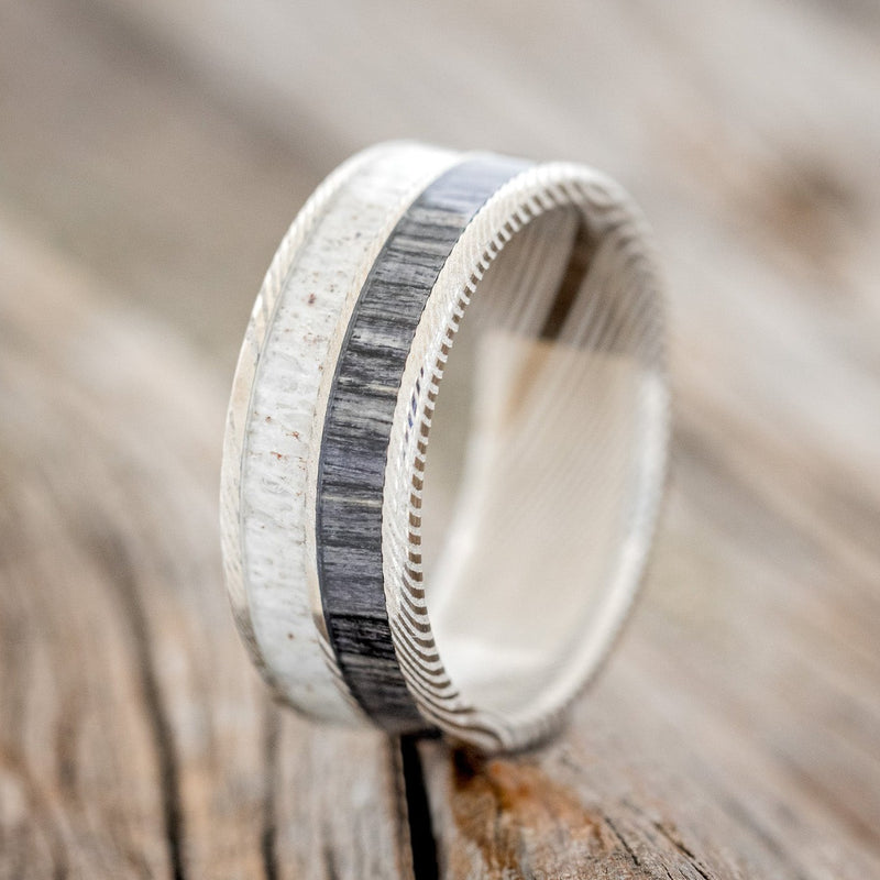 Shown here is "Dyad", a custom, handcrafted men's wedding ring featuring 2 channels with grey birch wood and antler inlays, shown here on an etched Damascus Steel band, upright facing left. Additional inlay options are available upon request.