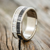 Shown here is "Dyad", a custom, handcrafted men's wedding ring featuring 2 channels with grey birch wood and antler inlays, shown here on a titanium band, upright facing left. Additional inlay options are available upon request.