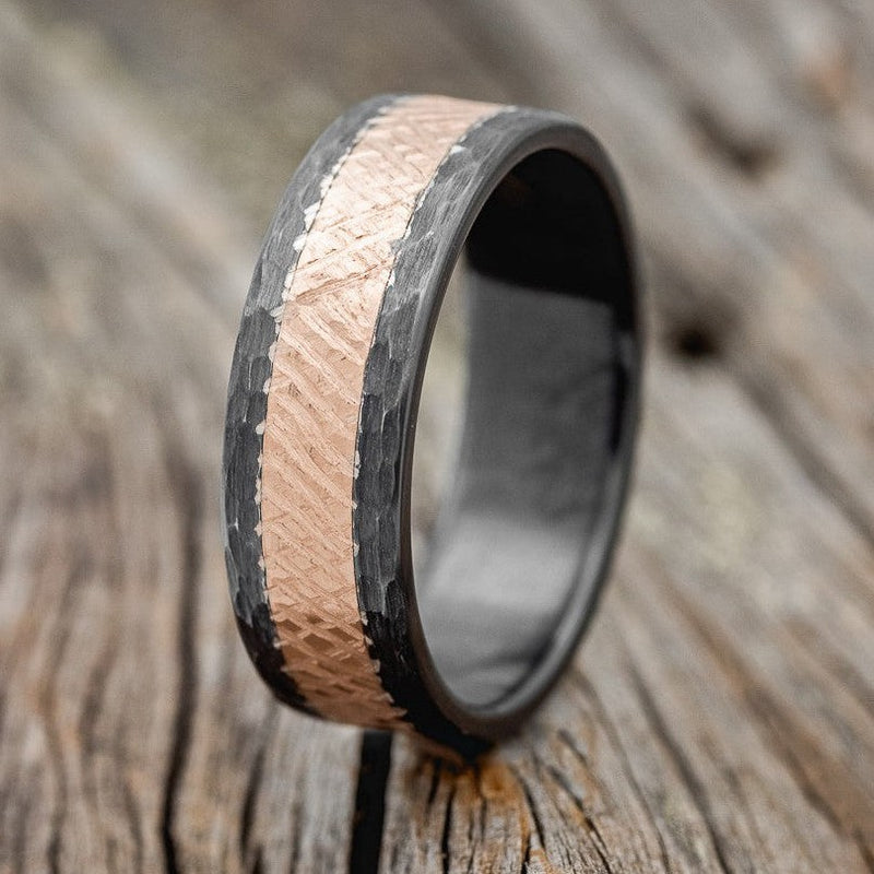 Shown here is a custom, handcrafted men's wedding ring featuring a solid metal band with a crosshatched finish 14K rose gold inlay, shown here in hammered black zirconium band, upright facing left. Additional inlay options are available upon request.