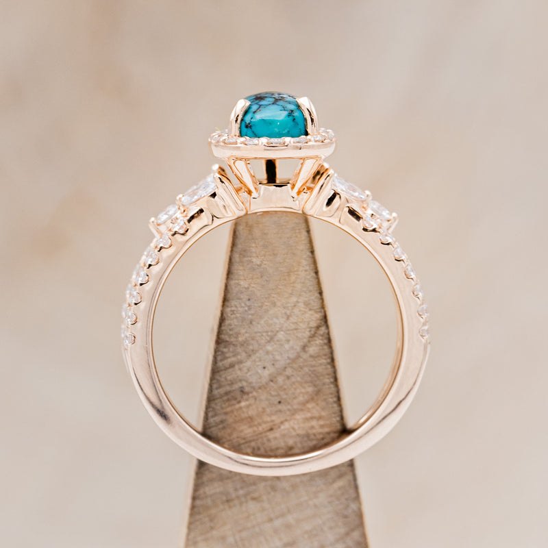 Shown here is "Loretta", a split shank-style turquoise women's engagement ring with a diamond halo and leaf-shape diamond accents, side view on stand. Many other center stone options are available upon request.