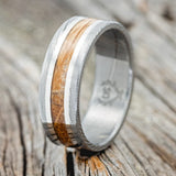 Shown here is "Kalder", a custom, handcrafted men's wedding ring featuring a Damascus Steel band with a whiskey barrel oak inlay and two 14K white gold inlays, upright facing left. Additional inlay options are available upon request.