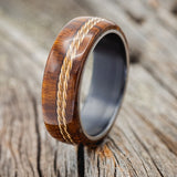 Shown here is "Remmy", a custom, handcrafted men's wedding ring featuring ironwood overlay and an offset twisted gold wire inlay, shown here on a black zirconium band, upright facing left. Additional inlay options are available upon request.