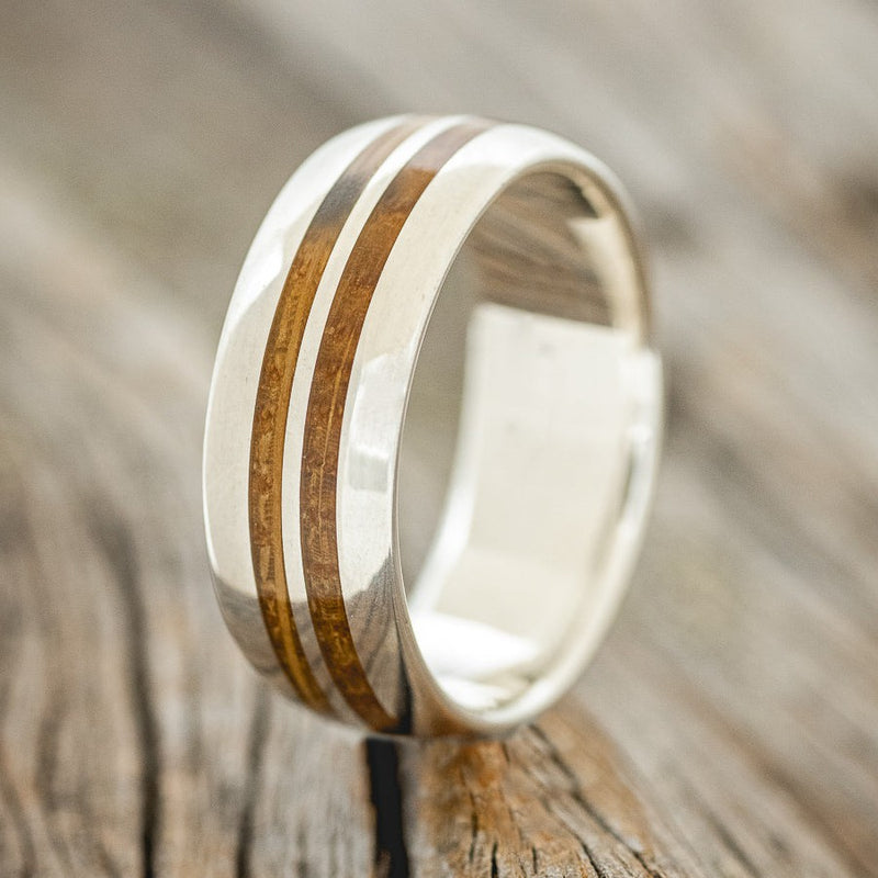 Shown here is "Silas", a handcrafted men's wedding ring featuring two whiskey barrel wood inlays on a domed profile silver band, upright facing left. Additional inlay options are available upon request.
