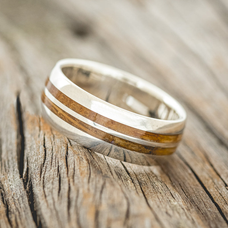 Shown here is "Silas", a handcrafted men's wedding ring featuring two whiskey barrel wood inlays on a domed profile silver band, tilted left. Additional inlay options are available upon request.