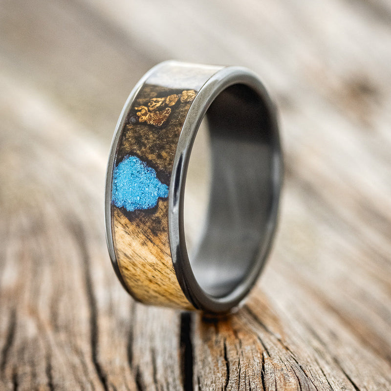 Shown here is "Rainier", a custom, handcrafted men's wedding ring featuring buckeye burl wood, with hand-crushed turquoise and gold nuggets filling the knots and burl holes, on a fire-treated black zirconium band, upright facing left. Additional inlay options are available upon request.