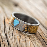 Shown here is "Rainier", a custom, handcrafted men's wedding ring featuring buckeye burl wood, with hand-crushed turquoise and gold nuggets filling the knots and burl holes, tilted left.