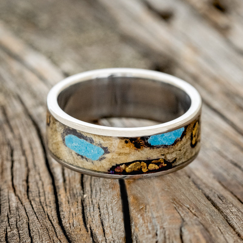 Shown here is "Rainier", a custom, handcrafted men's wedding ring featuring buckeye burl wood, with hand-crushed turquoise and gold nuggets filling the knots and burl holes, laying flat. Additional inlay options are available upon request.