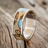 Shown here is "Rainier", a custom, handcrafted men's wedding ring featuring buckeye burl wood, with hand-crushed turquoise and gold nuggets filling the knots and burl holes, upright facing left. Additional inlay options are available upon request.