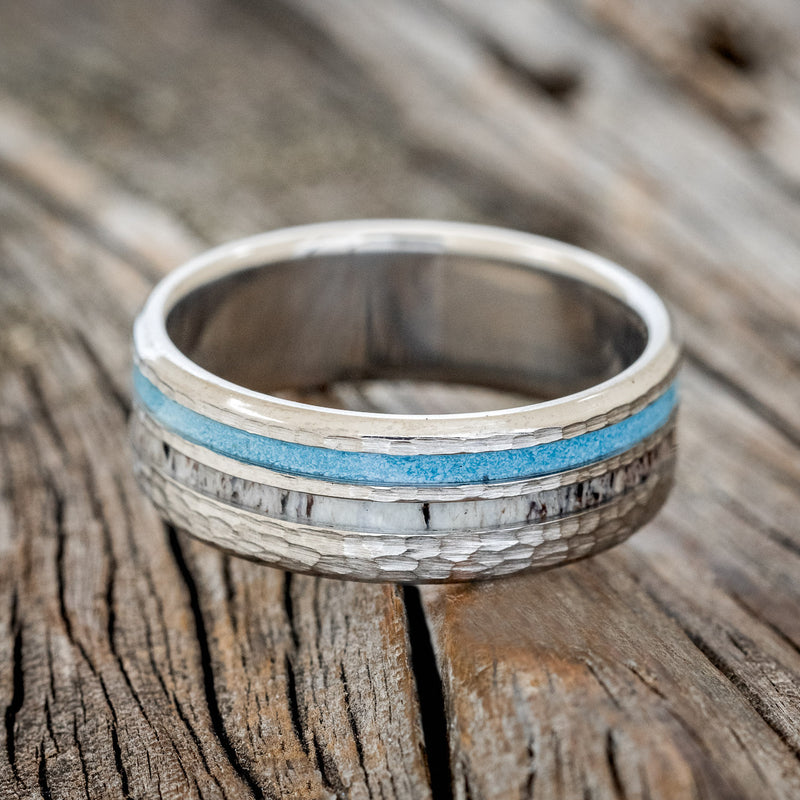 Shown here is "Cosmo", a custom, handcrafted men's wedding ring featuring elk antler and hand-crushed turquoise inlays on a hammered band, laying flat.