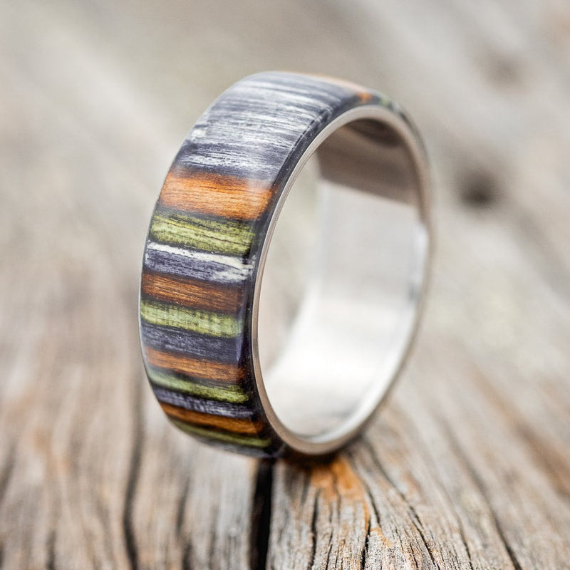 Shown here is "Haven", a custom, handcrafted men's wedding ring featuring birch wood overlay that has been dyed green, grey, and brown, upright facing left. Additional overlay options are available upon request.