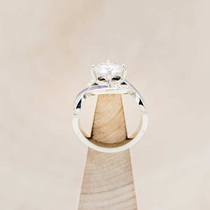 Shown here is "Helix", an infinity-style round cut moissanite women's engagement ring with diamond accents and sleepy lavender opal inlays, side view on stand. Many other center stone options are available upon request.