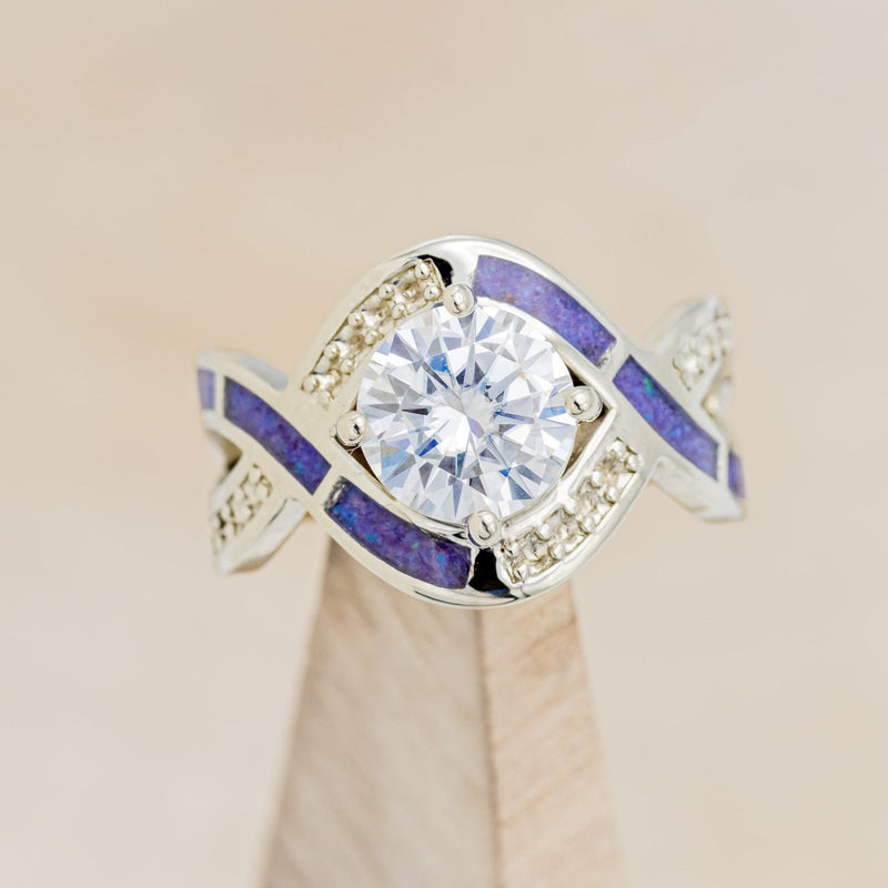 Shown here is "Helix", an infinity-style round cut moissanite women's engagement ring with diamond accents and sleepy lavender opal inlays, on stand front facing. Many other center stone options are available upon request. 
