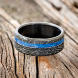 Shown here is "Vertigo", a custom, handcrafted men's wedding ring featuring a blue opal inlay on a hammered, fire-treated black zirconium band, laying flat. Additional inlay options are available upon request.