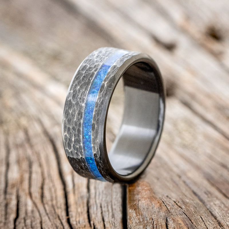 Shown here is "Vertigo", a handcrafted men's wedding ring featuring a blue opal inlay with a hammered, fire-treated black zirconium band, upright facing left.