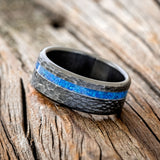 Shown here is "Vertigo", a custom, handcrafted men's wedding ring featuring a blue opal inlay on a hammered, fire-treated black zirconium band, tilted left. Additional inlay options are available upon request.
