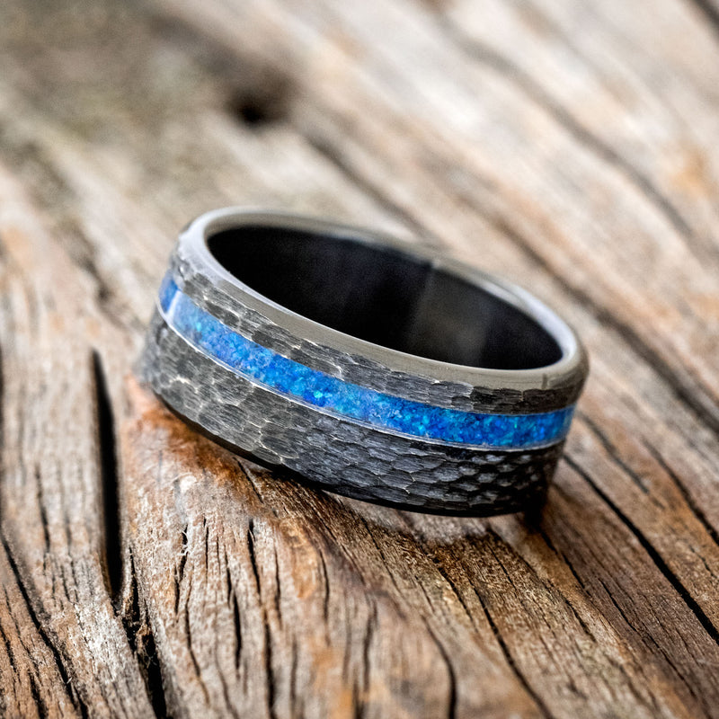 Shown here is "Vertigo", a handcrafted men's wedding ring featuring a blue opal inlay with a hammered, fire-treated black zirconium band, tilted left.