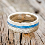 Shown here is "Vertigo", a custom, handcrafted men's wedding ring featuring a blue opal inlay on a hammered band, laying flat. Additional inlay options are available upon request.