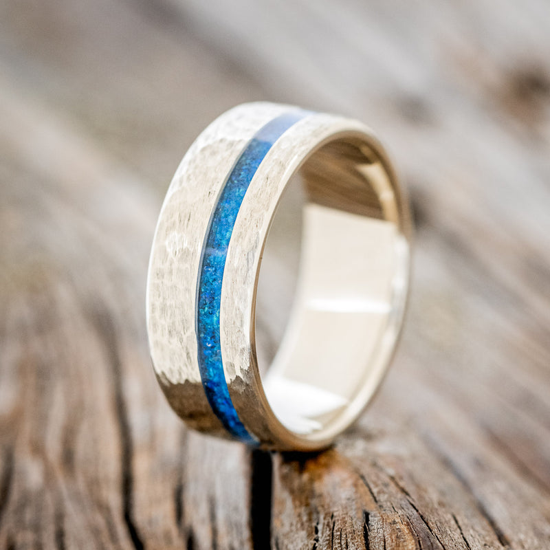 Shown here is "Vertigo", a custom, handcrafted men's wedding ring featuring a blue opal inlay on a hammered band, upright facing left. Additional inlay options are available upon request.