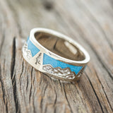 Shown here is "Summit", a custom, handcrafted men's wedding ring featuring a mountain range engraving with turquoise inlays, tilted left. Additional inlay options are available upon request.