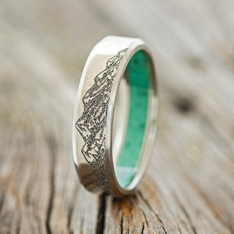 Shown here is a handcrafted men's wedding ring with a malachite-lined band and a mountain engraving, upright facing left. Additional inlay options are available upon request.