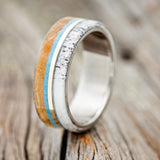 Shown here is "Banner", a custom, handcrafted men's wedding ring featuring a thin 14K yellow gold inlay set between whiskey barrel oak, turquoise, and antler shown here on a titanium band, upright facing left. Additional inlay options are available upon request.