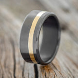 Shown here is "Vertigo", a custom, handcrafted men's wedding ring featuring a 14K yellow gold inlay on a fire-treated black zirconium band, upright facing left. Additional inlay options are available upon request.